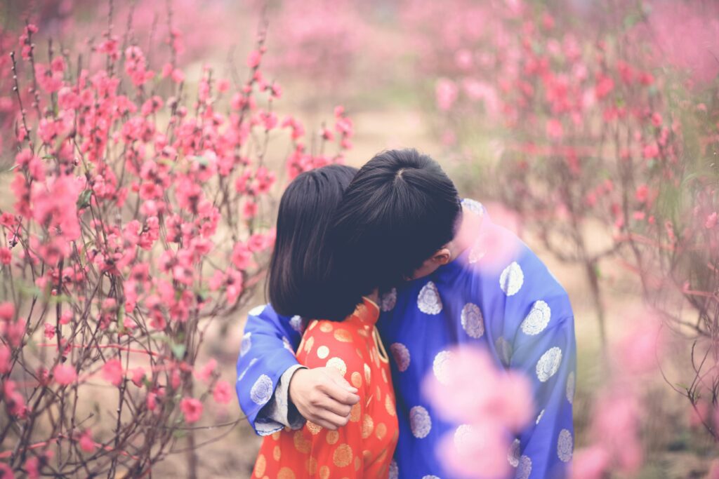 A couple standing in a field of flowers, leaning towards each other and appearing to smell the flowers.