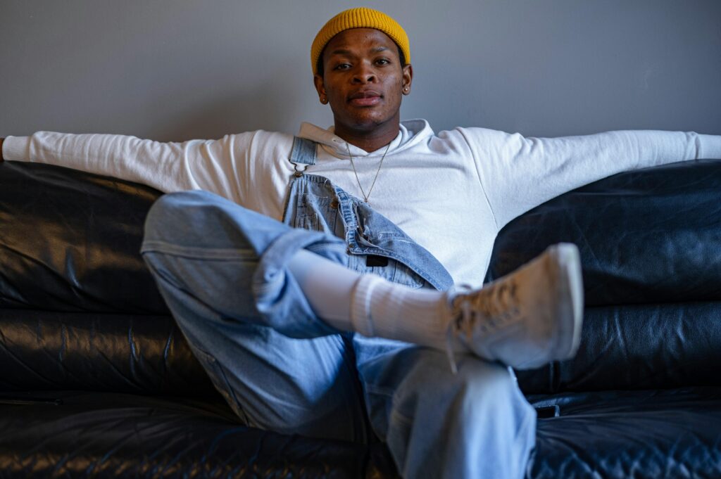 A confident Black man sitting on a sofa, wearing a hat stylishly.