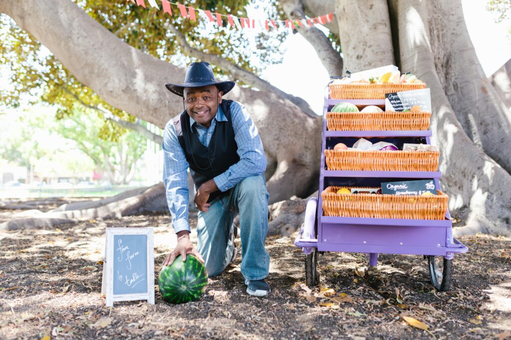 A man sitting at his stall, wearing a hat and smiling warmly.