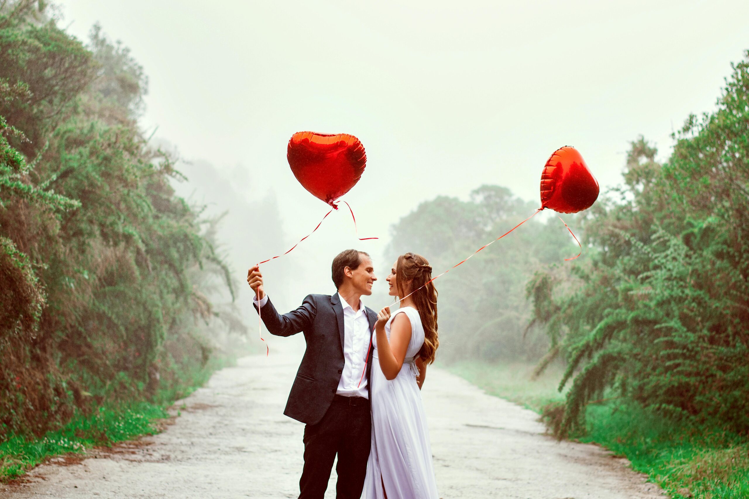 A young couple in a garden, both holding red heart balloons, with the guy asking, 'What does it mean when a guy asks you to be his valentine?