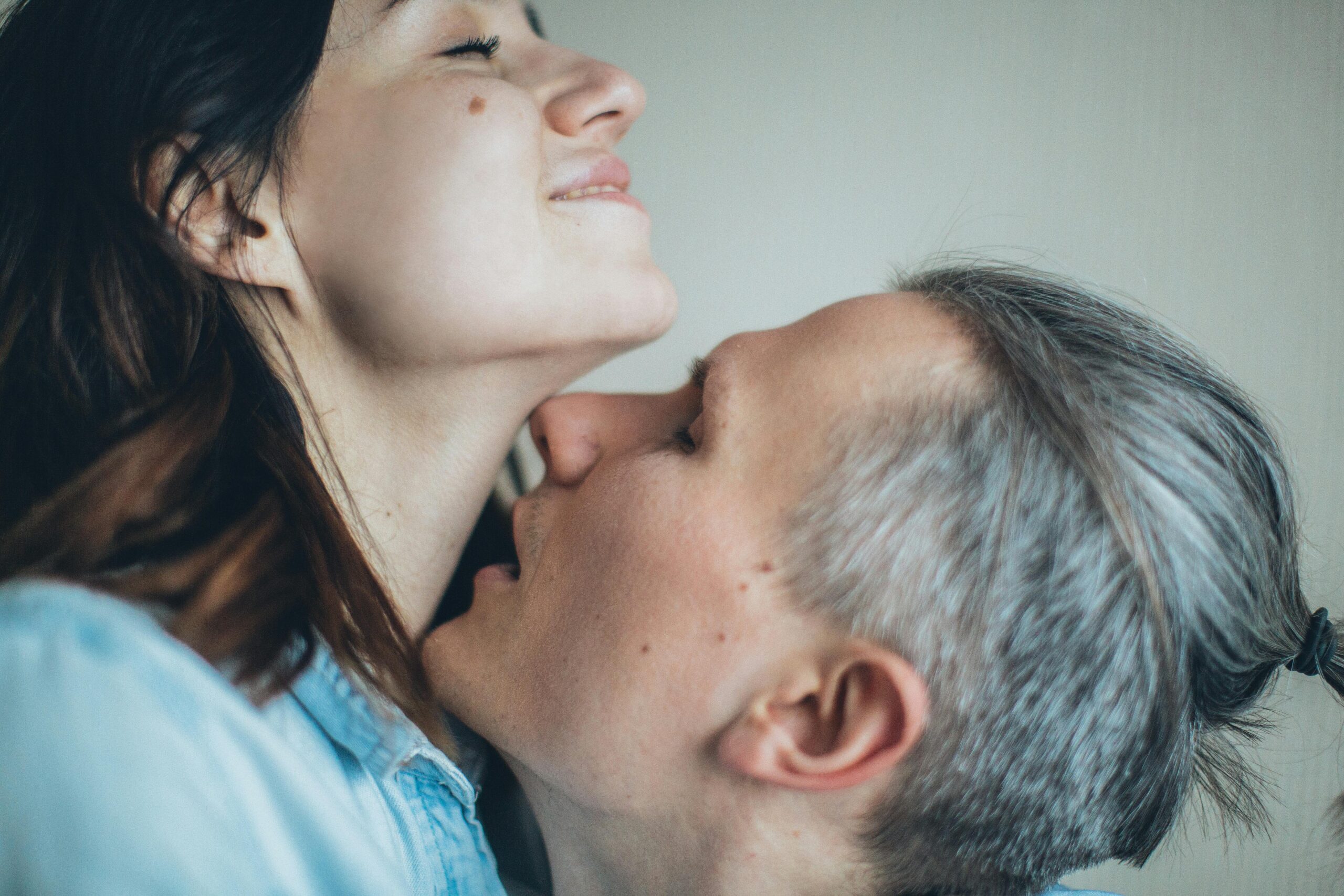 A couple standing in a room, with the man kissing his partner's neck, wondering about the meaning behind this intimate gesture.