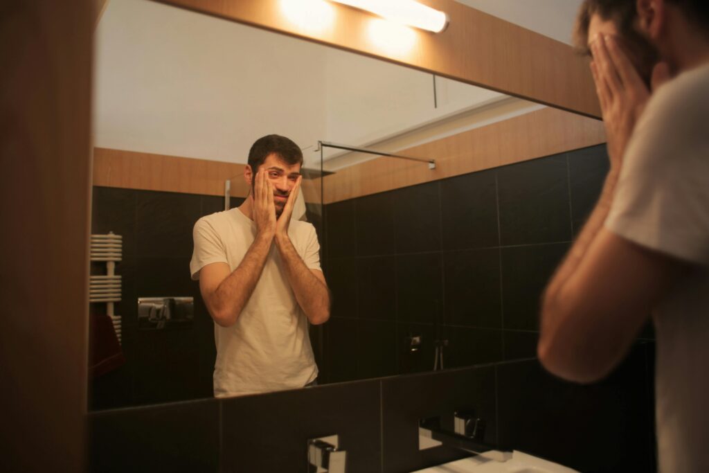 A man standing in front of a mirror, looking remorseful and sad after insulting his girlfriend.